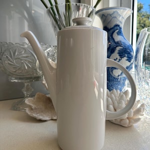 May include: A white ceramic teapot with a tall, cylindrical body and a spout. The teapot has a simple, modern design.