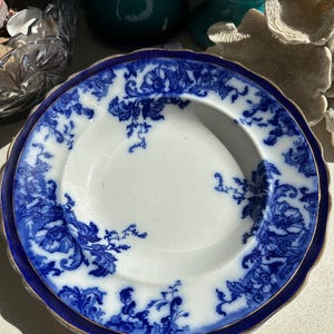 Puede incluir: Plato blanco con estampado floral azul y borde dorado. El plato tiene un borde festoneado y un borde azul oscuro. El dise&ntilde;o floral es un patr&oacute;n cl&aacute;sico y detallado. Una pieza de vajilla vintage.