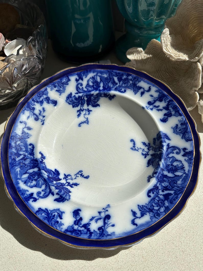 Puede incluir: Plato blanco con estampado floral azul y borde dorado. El plato tiene un borde festoneado y un borde azul oscuro. El dise&ntilde;o floral es un patr&oacute;n cl&aacute;sico y detallado. Una pieza de vajilla vintage.