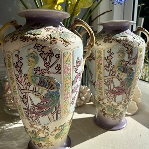 May include: Two ornate, vintage vases with a cream-colored base and lavender accents. Each vase features a detailed peacock design with colorful plumage and gold handles. The vases are decorated with floral and branch motifs.