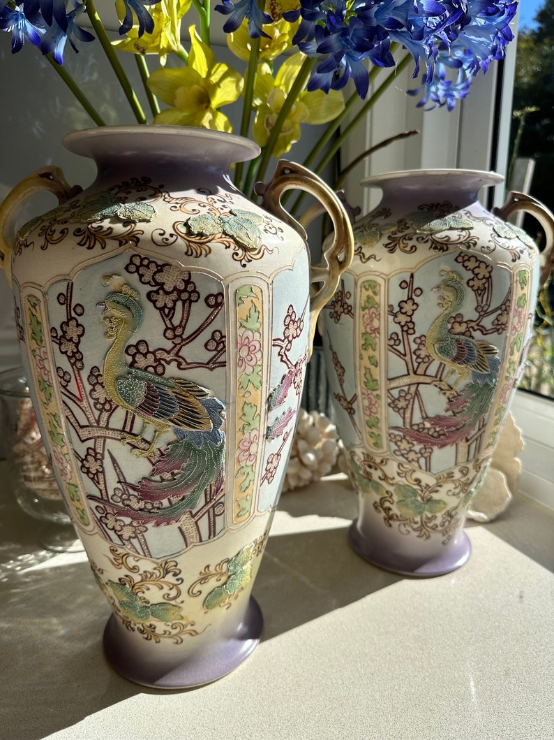 May include: Two ornate, vintage vases with a cream-colored base and lavender accents. Each vase features a detailed peacock design with colorful plumage and gold handles. The vases are decorated with floral and branch motifs.