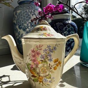 May include: A cream-coloured ceramic teapot featuring a floral pattern with pink, blue and yellow blossoms. The teapot has a matching lid, a curved handle, a pointed spout and a scalloped base with gold-toned trim along the edges and handle.