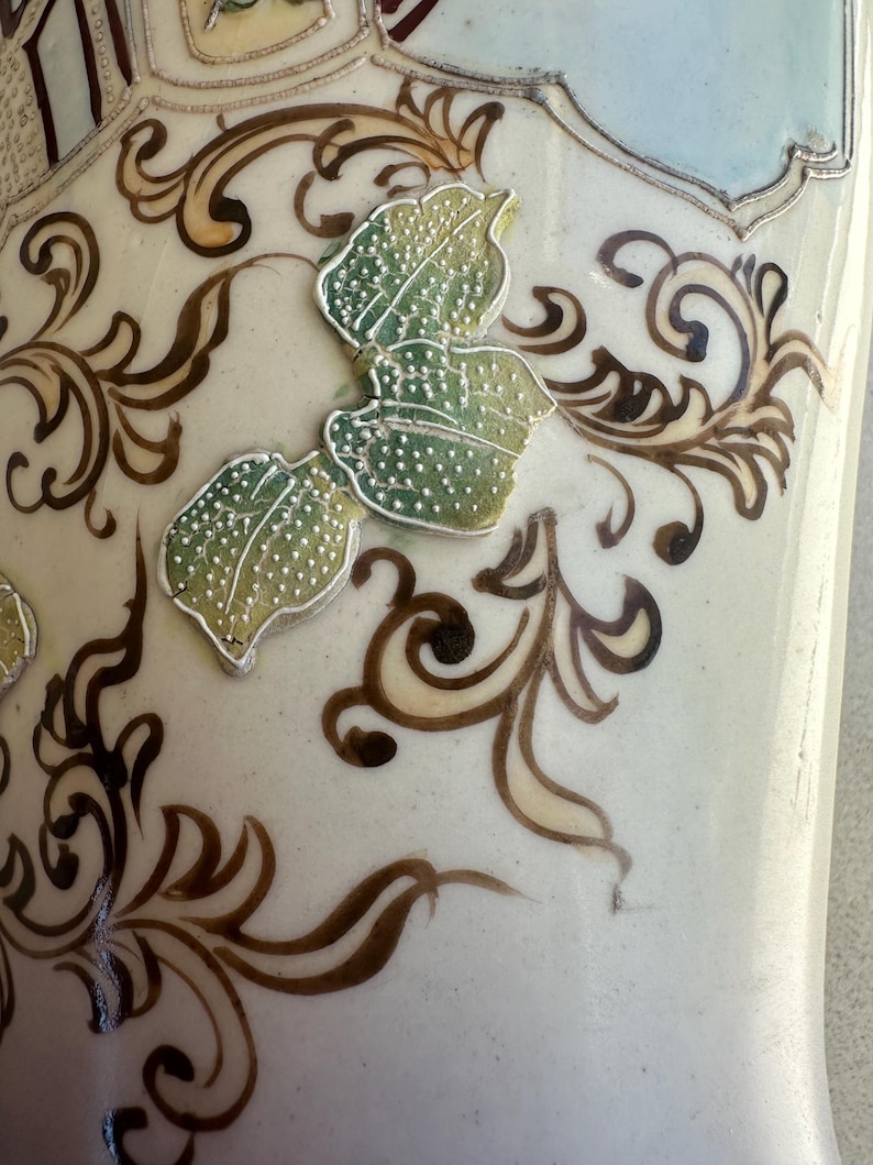 May include: Close-up of a ceramic vase with a cream-colored base. The vase features brown and gold scrollwork and three green leaves with silver outlines. The top of the vase has a light blue and maroon design.