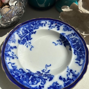 Puede incluir: Un plato antiguo blanco y azul con un dise&ntilde;o floral. El plato tiene un borde festoneado y un ribete dorado. El patr&oacute;n floral azul se encuentra sobre un fondo blanco. El plato es probablemente vintage o antiguo.