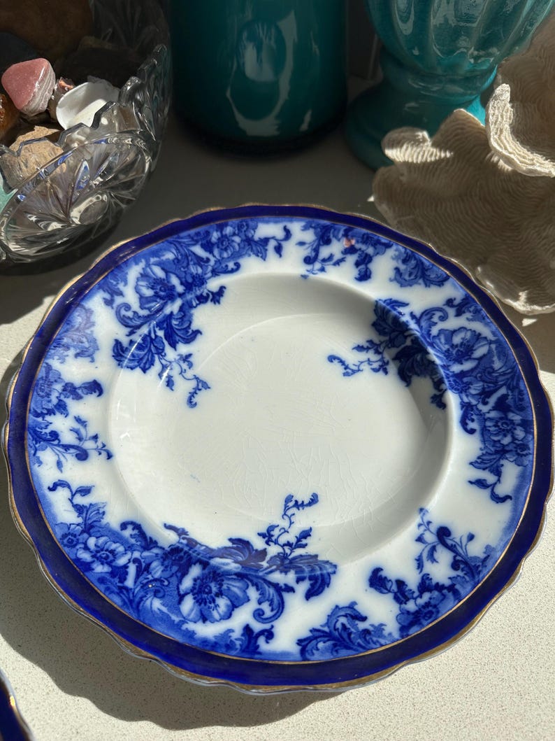 Puede incluir: Un plato antiguo blanco y azul con un dise&ntilde;o floral. El plato tiene un borde festoneado y un ribete dorado. El patr&oacute;n floral azul se encuentra sobre un fondo blanco. El plato es probablemente vintage o antiguo.