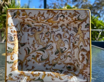 May include: A square ceramic dish with a yellow border and a white background. The dish is decorated with a floral pattern in shades of gold, green, and brown. There are four cherub figures in the center of the dish.