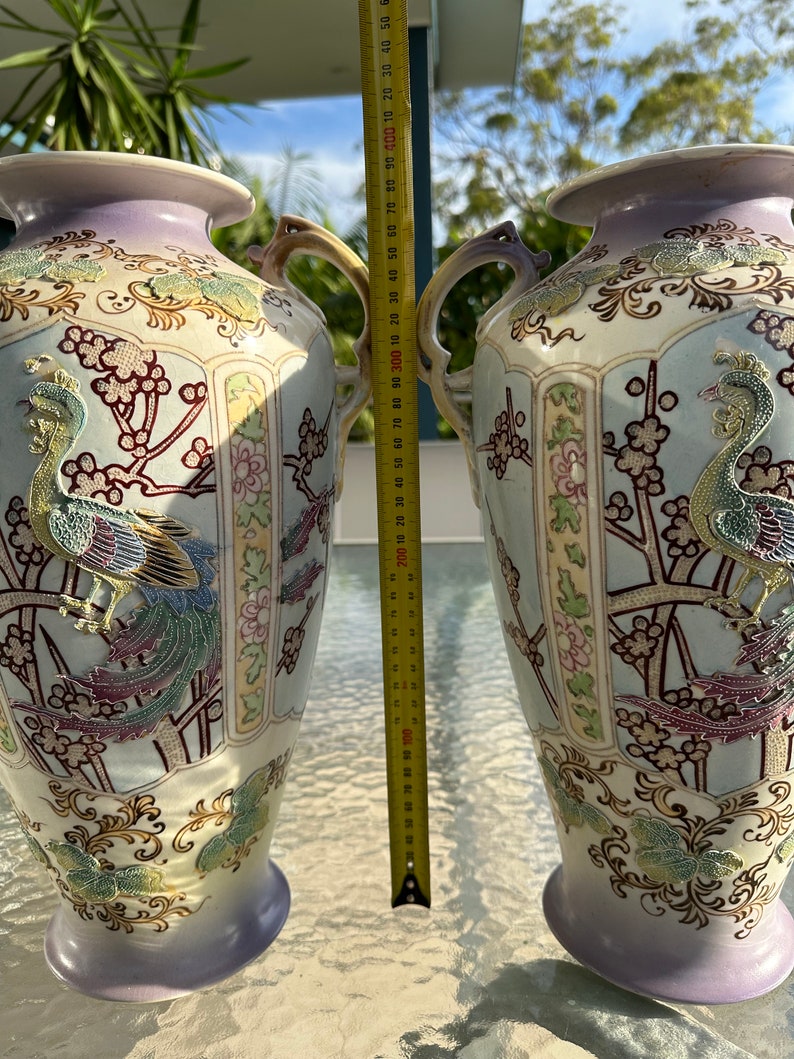 May include: Two ornate ceramic vases with handles, featuring a peacock design in gold, teal, and purple. The vases are approximately 16 inches tall, with a light blue and lavender color scheme, and floral accents.