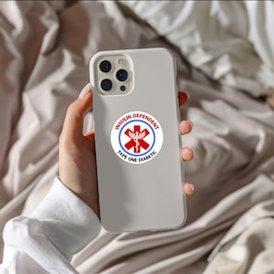 Puede incluir: Funda de teléfono gris claro con una pegatina circular que dice "INSULIN DEPENDENT TYPE ONE DIABETIC". La pegatina presenta un símbolo médico rojo y texto en un diseño circular. La funda del teléfono se sostiene en una mano.