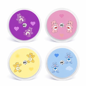 May include: Four circular decorative items with Care Bears designs. Each has a different coloured background: purple, pink, yellow, and blue. Each design includes Care Bears, hearts, a football, a sun, and clouds.