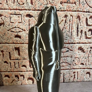 Może przedstawiać: Srebrna, trójwymiarowa rzeźba postaci ludzkiej stojącej z ramionami opuszczonymi wzdłuż ciała. Rzeźba ma gładkie, metaliczne wykończenie i stoi na jasnoszarym podłożu.