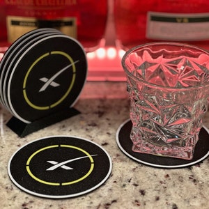 Peut inclure: Quatre sous-verres noirs avec un logo SpaceX blanc et un anneau jaune autour. Les sous-verres sont empilés les uns sur les autres. Un verre à facettes repose sur un sous-verre noir.