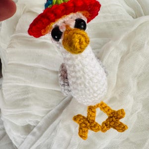 May include: A handmade crochet bird with a red, yellow, blue, and green hat. The bird has a yellow beak and feet, and black eyes. The bird is white and gray, and is hanging from a purple string.