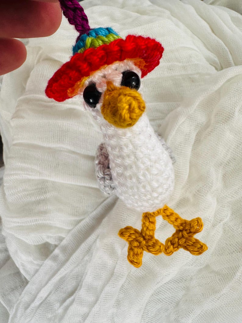 May include: A handmade crochet bird with a red, yellow, blue, and green hat. The bird has a yellow beak and feet, and black eyes. The bird is white and gray, and is hanging from a purple string.