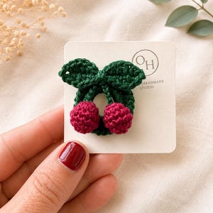 May include: A handmade crochet cherry hair clip. The clip features a green leaf design with two red cherries. The hair clip is on a white card with the text "OH HANDMADE STUDIO".