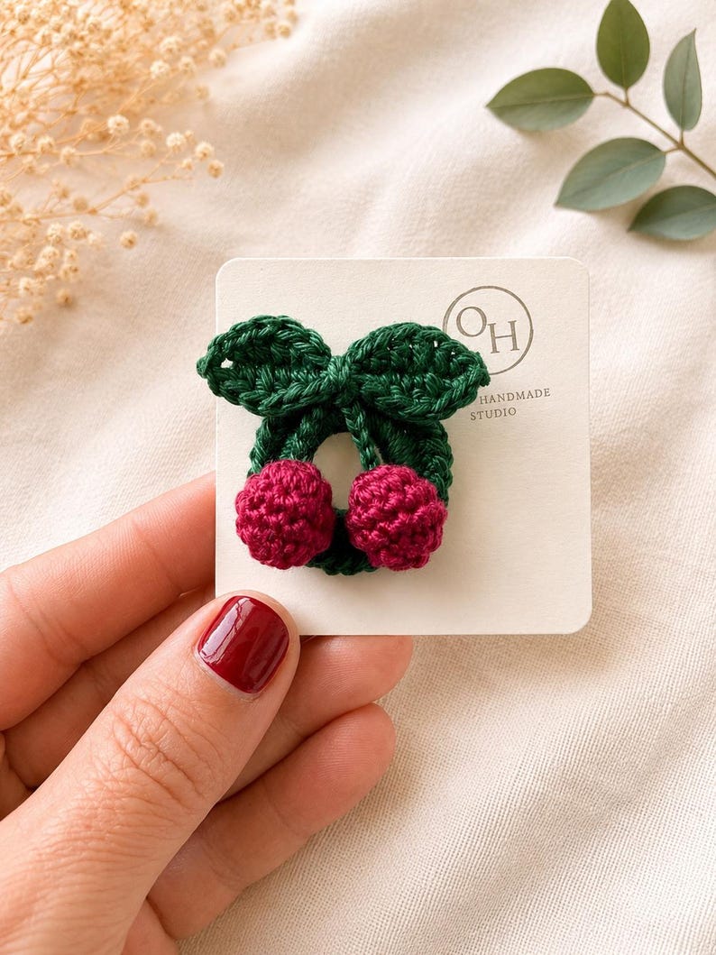 May include: A handmade crochet cherry hair clip. The clip features a green leaf design with two red cherries. The hair clip is on a white card with the text "OH HANDMADE STUDIO".