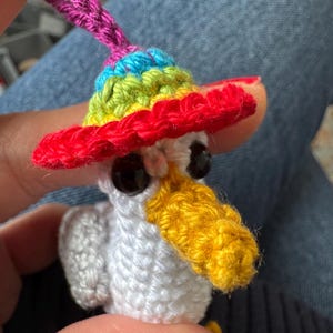 May include: A handmade crochet bird with a rainbow-colored sombrero. The bird has a white body, black eyes, and a yellow beak. A pink loop is attached to the top of the hat. The bird is a whimsical, colorful creation.