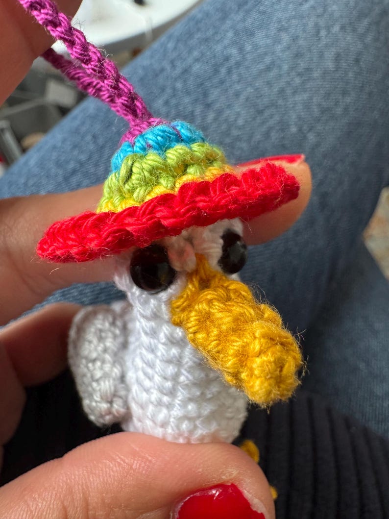 May include: A handmade crochet bird with a rainbow-colored sombrero. The bird has a white body, black eyes, and a yellow beak. A pink loop is attached to the top of the hat. The bird is a whimsical, colorful creation.