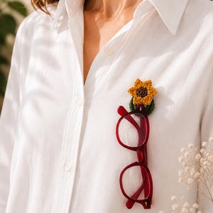May include: A white shirt with a crocheted sunflower pin securing red eyeglasses. The sunflower pin is yellow and brown, and the glasses have a dark red frame. The glasses are suspended from the pin, creating a unique accessory.