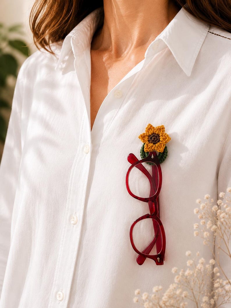 May include: A white shirt with a crocheted sunflower pin securing red eyeglasses. The sunflower pin is yellow and brown, and the glasses have a dark red frame. The glasses are suspended from the pin, creating a unique accessory.
