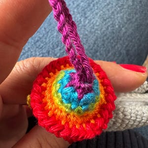 May include: A handmade, colorful crochet item with a rainbow spiral design. The central area is blue, surrounded by rings of green, yellow, orange, and red. A purple crochet cord extends from the top.