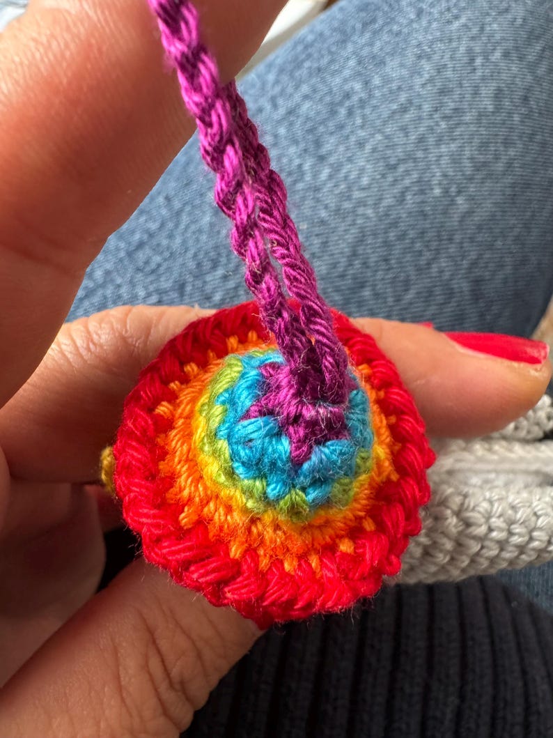May include: A handmade, colorful crochet item with a rainbow spiral design. The central area is blue, surrounded by rings of green, yellow, orange, and red. A purple crochet cord extends from the top.