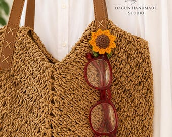 May include: A tan crocheted tote bag with brown faux leather straps and a small yellow and brown crocheted sunflower embellishment. A pair of dark red glasses hangs from the sunflower. The logo OH OZGUN HANDMADE STUDIO and the URL www.ozgunhandmade.co.uk are visible.