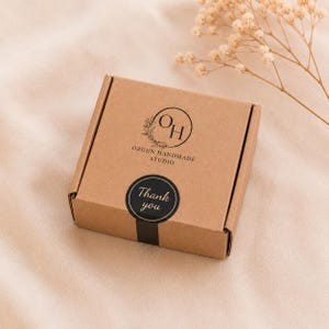 May include: A small, square, brown cardboard box with a black sticker that says "Thank you." The box has a logo that says "OH" inside a circle with a floral design and the text "OZGUN HANDMADE STUDIO."