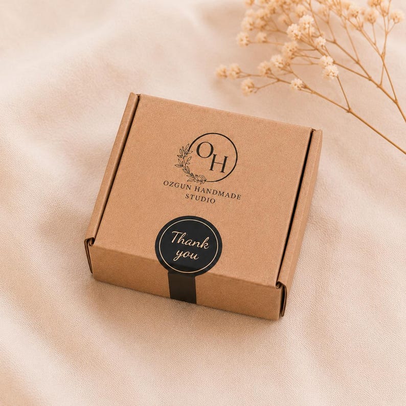 May include: A small, square, brown cardboard box with a black sticker that says "Thank you." The box has a logo that says "OH" inside a circle with a floral design and the text "OZGUN HANDMADE STUDIO."