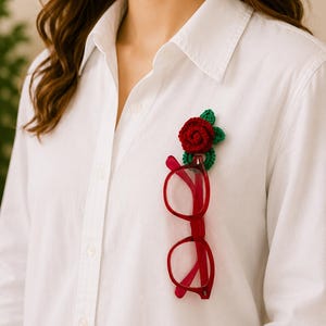 May include: A white shirt with a red-framed pair of spectacles clipped to a crocheted red rose and green leaves. The rose is attached to the shirt, creating a unique accessory.