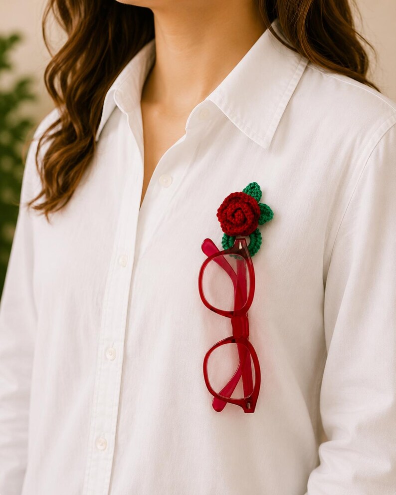 May include: A white shirt with a red-framed pair of spectacles clipped to a crocheted red rose and green leaves. The rose is attached to the shirt, creating a unique accessory.