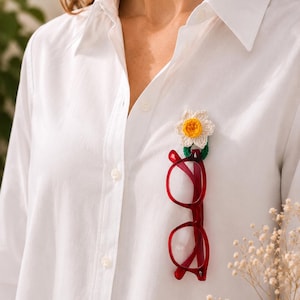 May include: A white shirt with a crochet flower brooch and red spectacles attached. The crochet flower has white petals and a yellow centre. The spectacles are a bright red colour.
