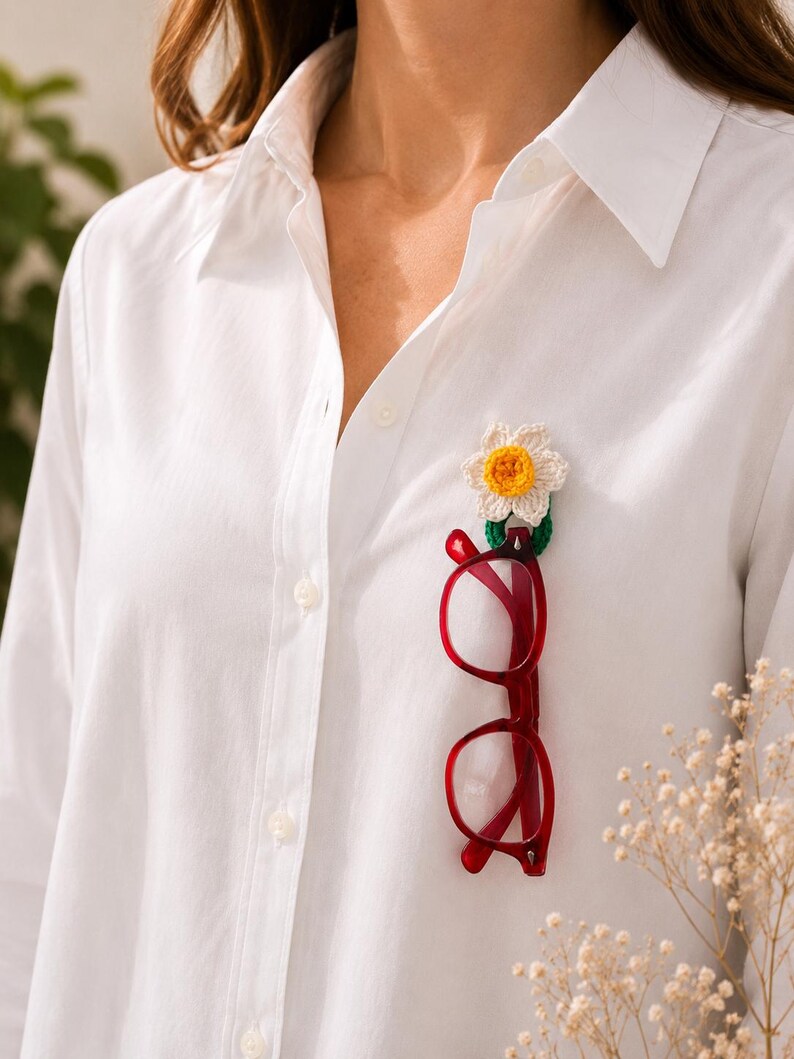 May include: A white shirt with a crochet flower brooch and red spectacles attached. The crochet flower has white petals and a yellow centre. The spectacles are a bright red colour.