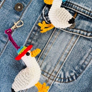 May include: Two crochet duck ornaments with hats, one with a rainbow brim and the other with a blue hat, are displayed on a denim jacket. The ducks have white bodies, yellow feet and beaks, and black accents.