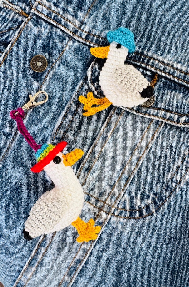 May include: Two crochet duck ornaments with hats, one with a rainbow brim and the other with a blue hat, are displayed on a denim jacket. The ducks have white bodies, yellow feet and beaks, and black accents.