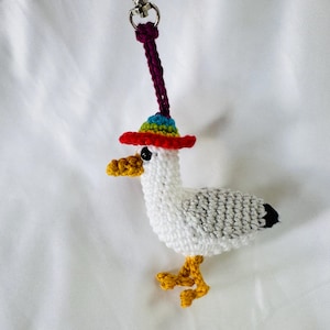 May include: A crocheted seagull keychain with a red, rainbow-brimmed hat. The seagull has white and gray body, yellow feet, and a yellow beak. A silver-colored clasp and purple cord are attached to the top of the hat.