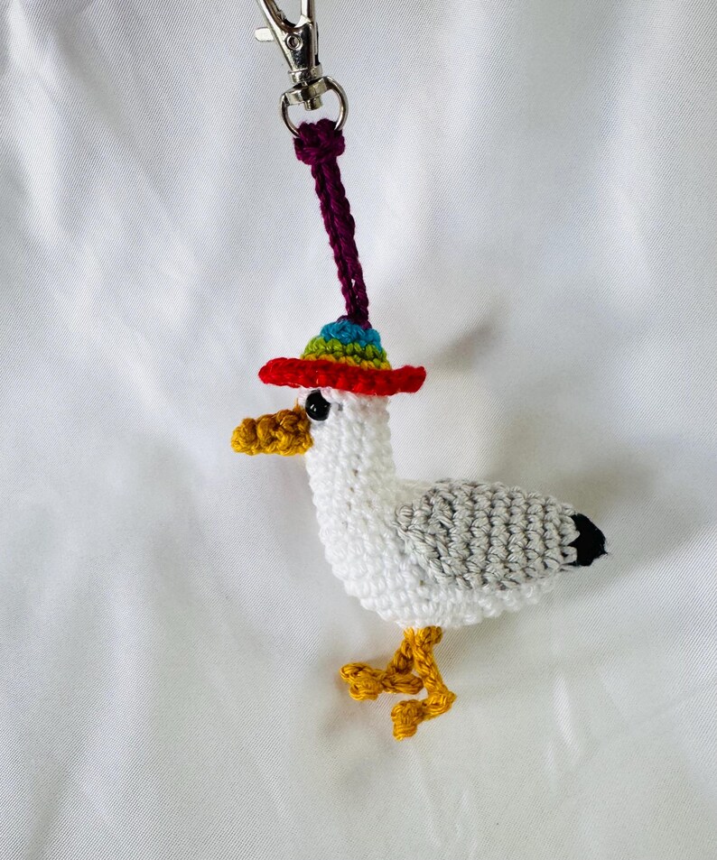 May include: A crocheted seagull keychain with a red, rainbow-brimmed hat. The seagull has white and gray body, yellow feet, and a yellow beak. A silver-colored clasp and purple cord are attached to the top of the hat.