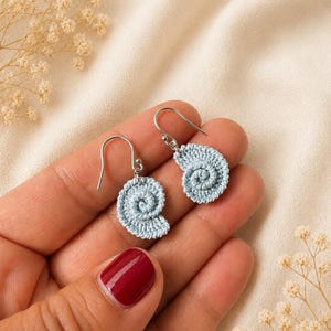 Peut inclure: Une paire de boucles d'oreilles en crochet bleu clair en forme de coquillage en spirale. Les boucles d'oreilles ont des crochets argentés et sont présentées sur un tissu crème. Bijou fait main avec un motif délicat et complexe.