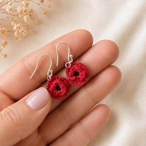 May include: Handmade crochet poppy earrings. The earrings have bright red flower petals with a black centre and silver-coloured ear wires. The earrings are held in a hand against a soft, neutral background.