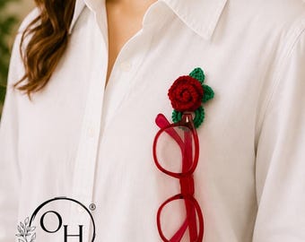 May include: A red crocheted rose brooch with green leaves attached to a white button-down shirt, functioning as a holder for a pair of red-framed spectacles. The brooch features a circular loop at the bottom to secure the glasses in place.