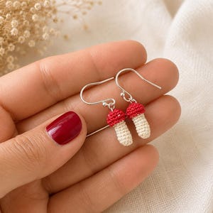 May include: A pair of handmade crochet mushroom earrings. Each earring features a red cap and a cream-colored stem, suspended from silver-colored ear wires. The earrings are small, detailed, and appear to be lightweight.