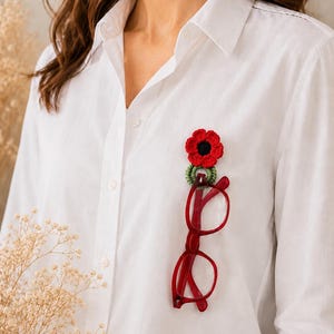 May include: A red crochet poppy flower brooch with a black centre and green detailing, alongside red-framed spectacles. The brooch and glasses are attached to a white button-down shirt, creating a unique accessory.