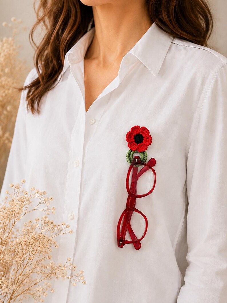 May include: A red crochet poppy flower brooch with a black centre and green detailing, alongside red-framed spectacles. The brooch and glasses are attached to a white button-down shirt, creating a unique accessory.