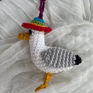 May include: A handmade crochet seagull keychain. The seagull is white with gray wings, a yellow beak and legs, and a black tail. It wears a colorful rainbow hat with a red brim. The keychain has a purple cord and a silver clasp.
