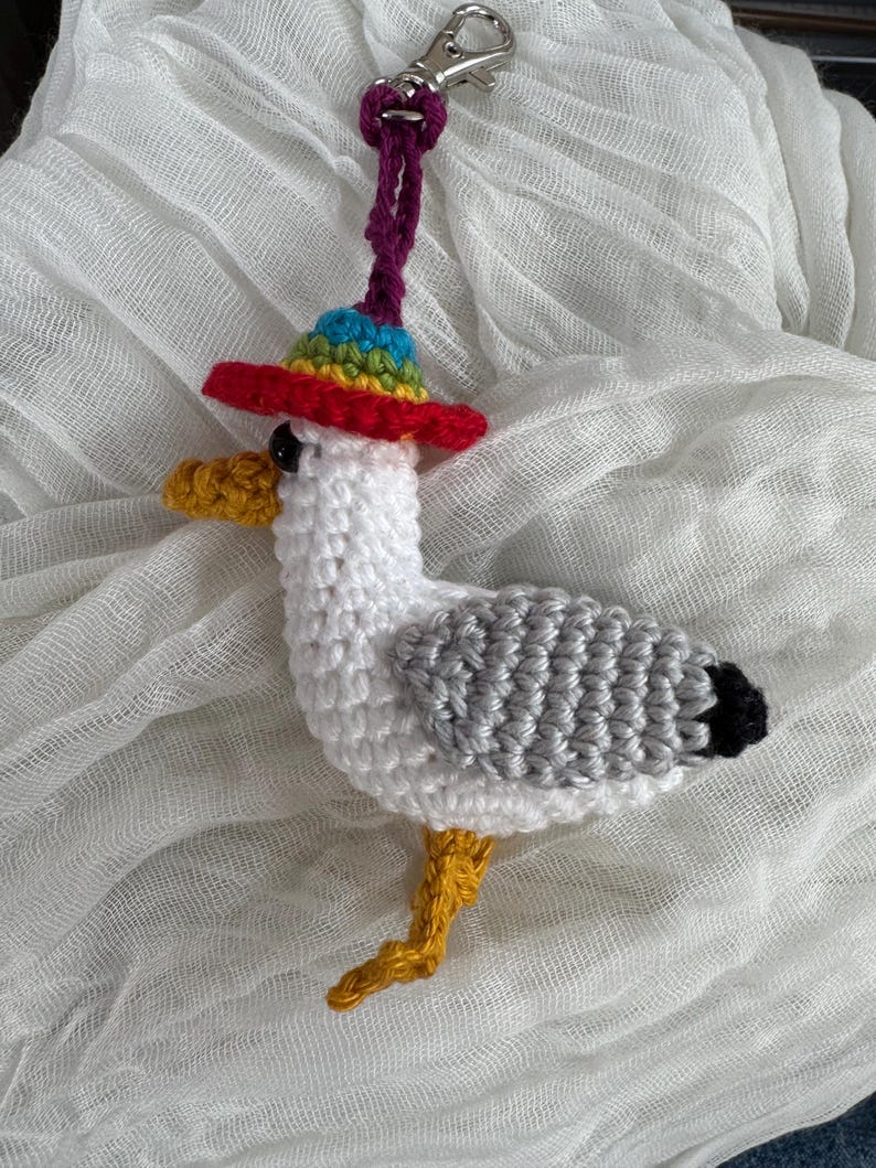 May include: A handmade crochet seagull keychain. The seagull is white with gray wings, a yellow beak and legs, and a black tail. It wears a colorful rainbow hat with a red brim. The keychain has a purple cord and a silver clasp.