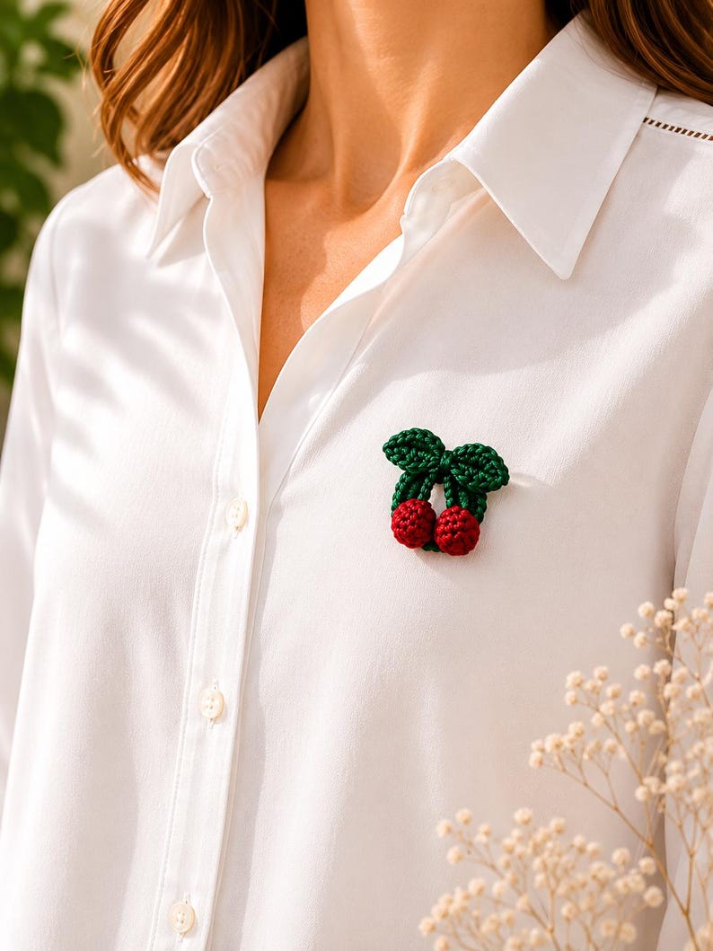 May include: A crochet cherry brooch in green and red, attached to a white button-down shirt. The brooch depicts two red cherries and green leaves. The shirt is buttoned and has a classic collar.