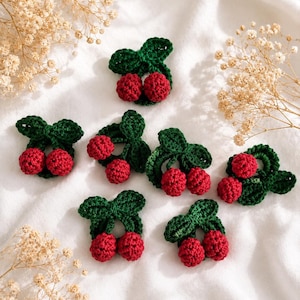 May include: A collection of handmade crochet cherry appliques. Each applique has two red cherries with green leaves and stems. The appliques are displayed on a white surface, with dried flowers in the background. Perfect for crafting and embellishments.