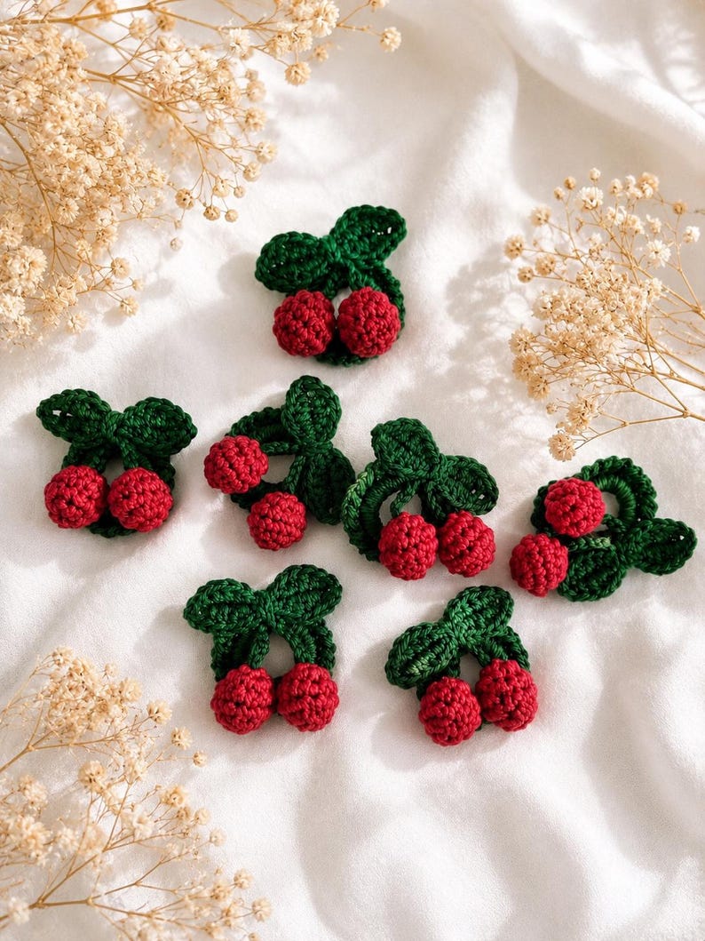 May include: A collection of handmade crochet cherry appliques. Each applique has two red cherries with green leaves and stems. The appliques are displayed on a white surface, with dried flowers in the background. Perfect for crafting and embellishments.