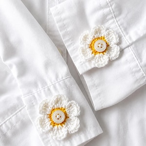 May include: A crisp white long-sleeved button-down shirt featuring two decorative crochet flower appliqués attached to the cuffs. Each flower has white petals and a yellow center, with a small white four-hole button secured in the middle.