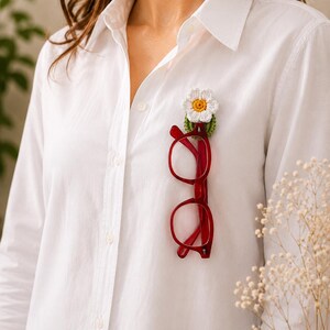 May include: A white shirt with a red-framed pair of glasses clipped to the front. A handmade crochet flower pin, featuring a white daisy and yellow centre, is attached to the shirt.