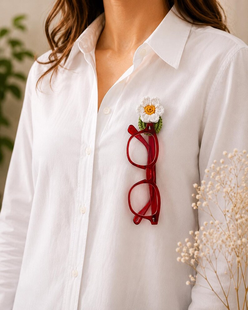 May include: A white shirt with a red-framed pair of glasses clipped to the front. A handmade crochet flower pin, featuring a white daisy and yellow centre, is attached to the shirt.